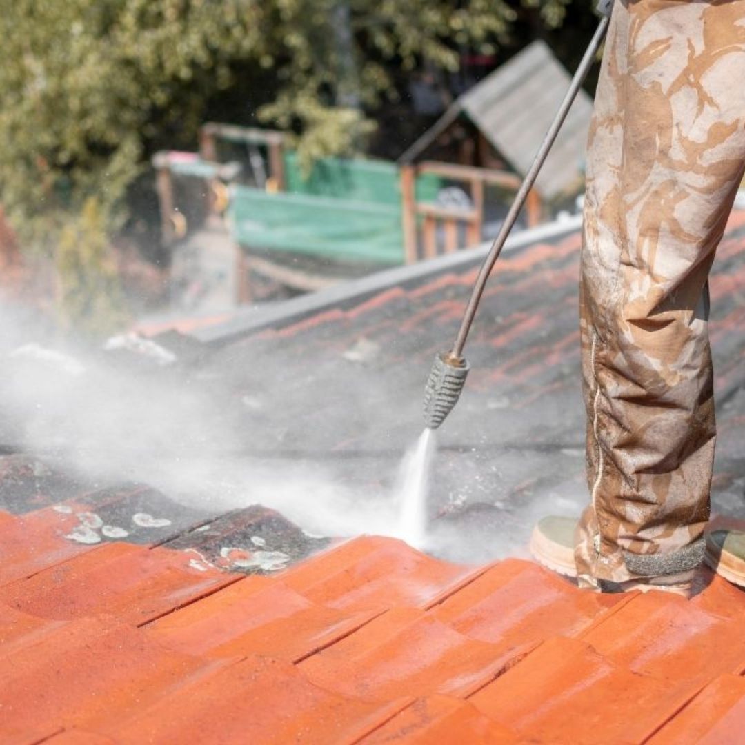 Technician washing terracotta roof tiles, revealing clean orange tile beneath dirt and algae buildup